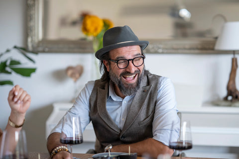 Ein lachender Mann sitzt an einem mit köstlichen Tapas gedeckten Tisch. Vor ihm stehen mehrere Gläser, während er mit zugekniffenen Augen herzhaft lacht und ein Wein-Tasting genießt. Die Atmosphäre wirkt gesellig und entspannt.