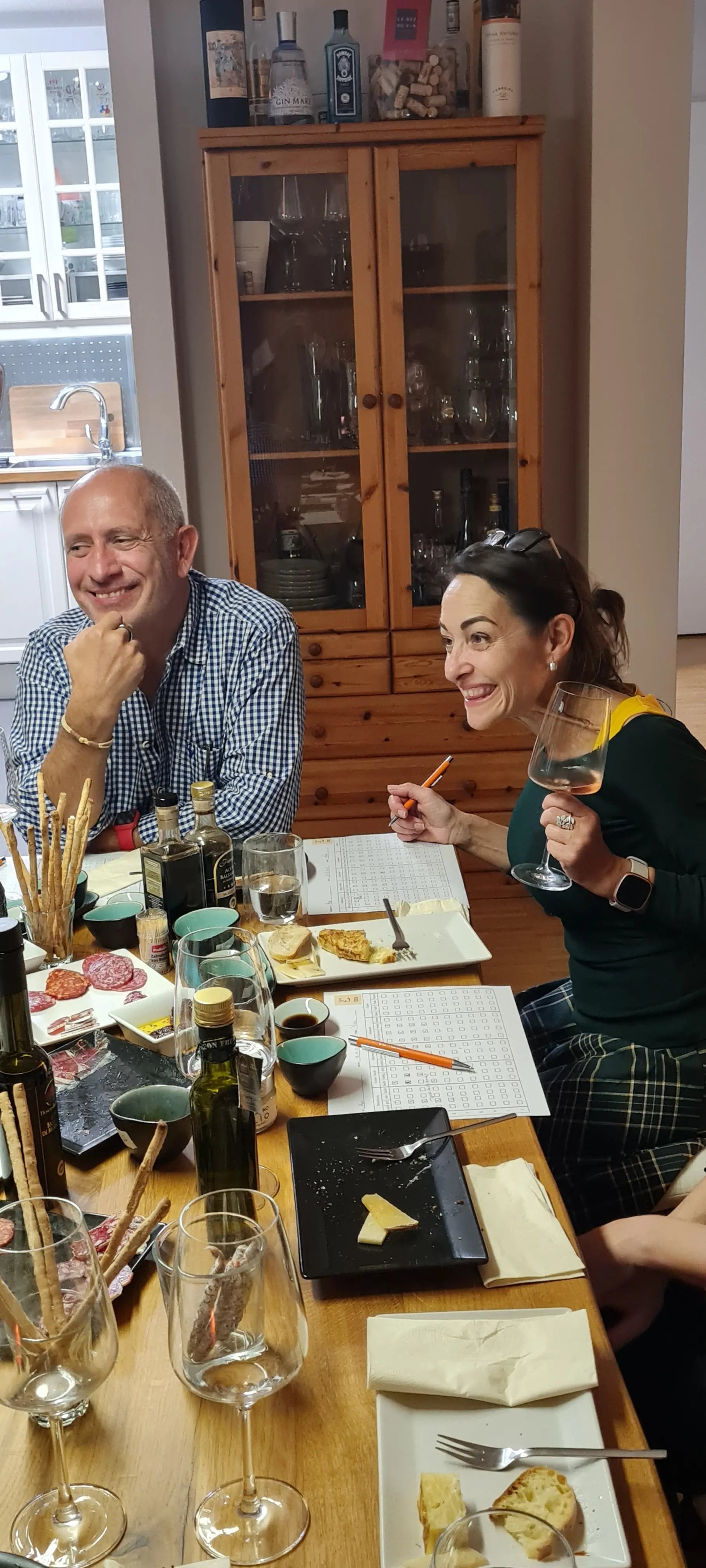 Eine Frau und ein Mann strahlen vor Freude sitzen am gedeckten Tisch zum Tasting zusammen. Die Frau hat ein Glas Rotwein in der einen Hand und in der anderen einen Kugelschreiber. Auf dem Tisch stehen vielen Tapas, Gläsern, Flaschen und Beurteilungszetteln. 